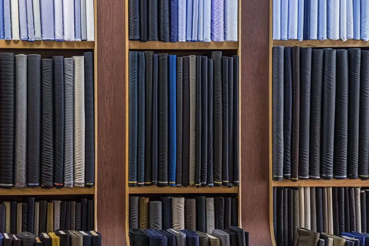 Wall of suiting rolls in navy, charcoal and grey with folded shirtings on the top shelf