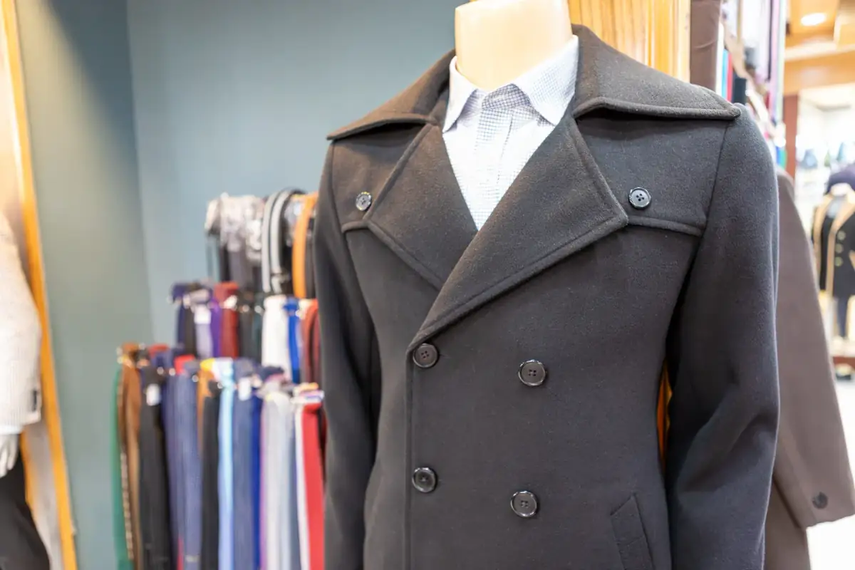 Charcoal double-breasted wool overcoat on a mannequin beside a rack of trouser lengths